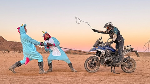 Zwei Fahrer des deutschen Männerteams in Einhorn-Kostümen ziehen an einem Abschleppseil, das an einer R 1300 GS befestigt ist. Der dritte Fahrer sitzt auf dem Motorrad und schwingt eine Peitsche.
